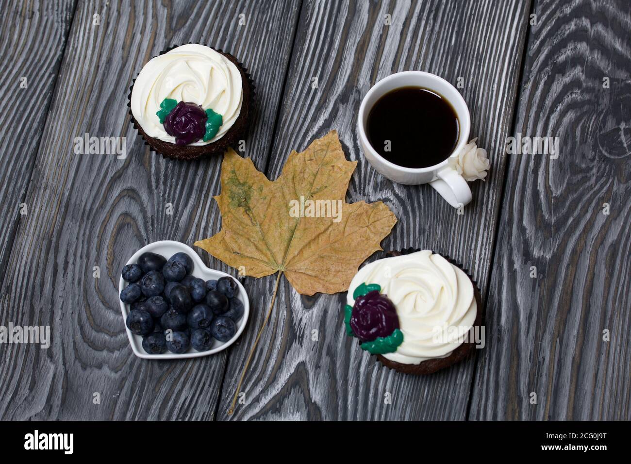 Muffin al cioccolato con fiore alla crema. Una tazza di caffè nero. Piatto con mirtilli. Foglia di acero essiccata. Si trova su tavole di pino dipinte in bianco e nero. Foto Stock
