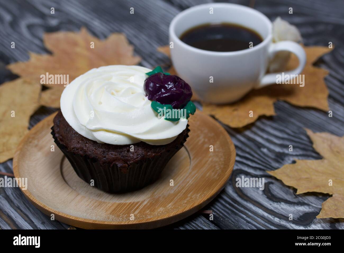 Muffin al cioccolato con fiore alla crema. Una tazza di caffè nero. Tra le foglie di acero essiccate. Giacciono su tavole di pino dipinte in bianco e nero. Foto Stock