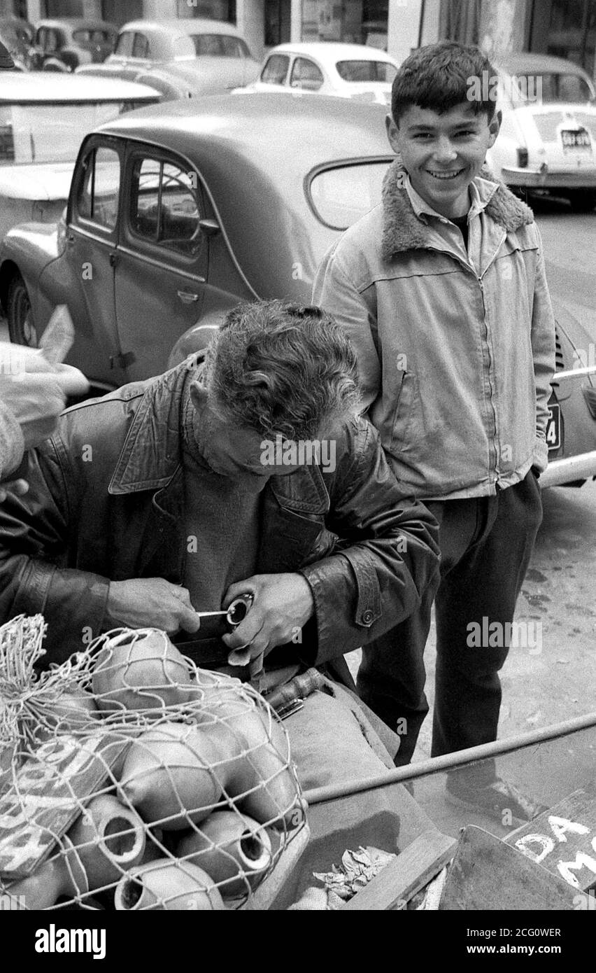 AJAXNETPHOTO. 1964. MONTEVIDEO, URUGUAY. - STREET CARVER - UOMO CHE INTAGLIA CALABASH GOURD CONCHIGLIE SU UNA STRADA DELLA CITTÀ MENTRE UN GIOVANE SORRIDE ALLA CAMERA.PHOTO:JONATHAN EASTLAND/AJAX REF:35646 24 71 Foto Stock