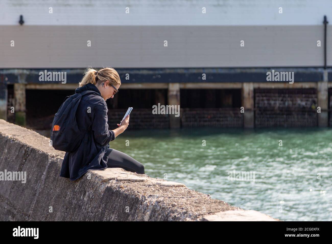 Una giovane donna seduta su un muro al mare guardando il suo telefono cellulare o cellulare Foto Stock
