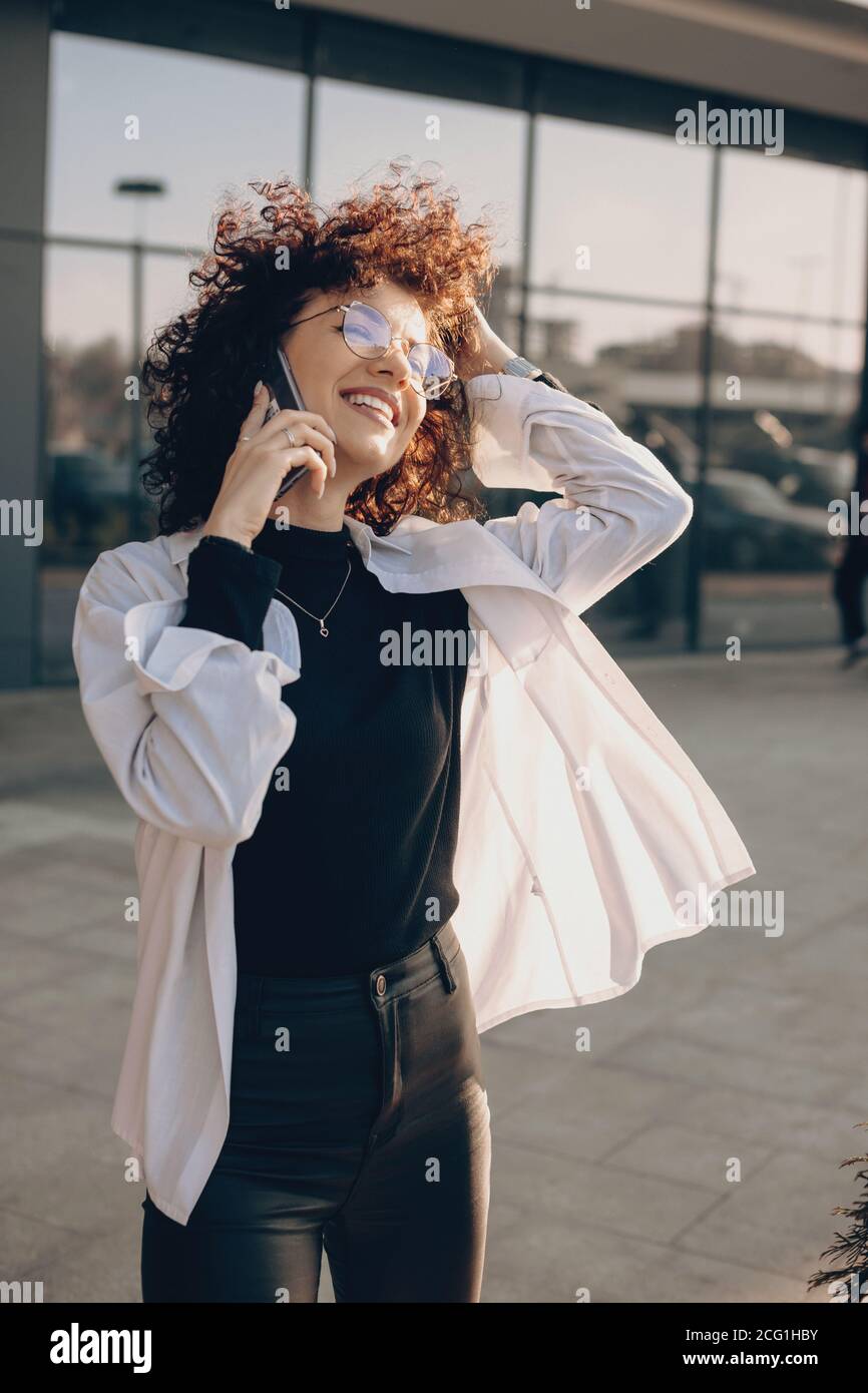 Felice ragazza d'affari con capelli ricci con occhiali che parlano telefona vicino al suo ufficio e sorridi al sole Foto Stock