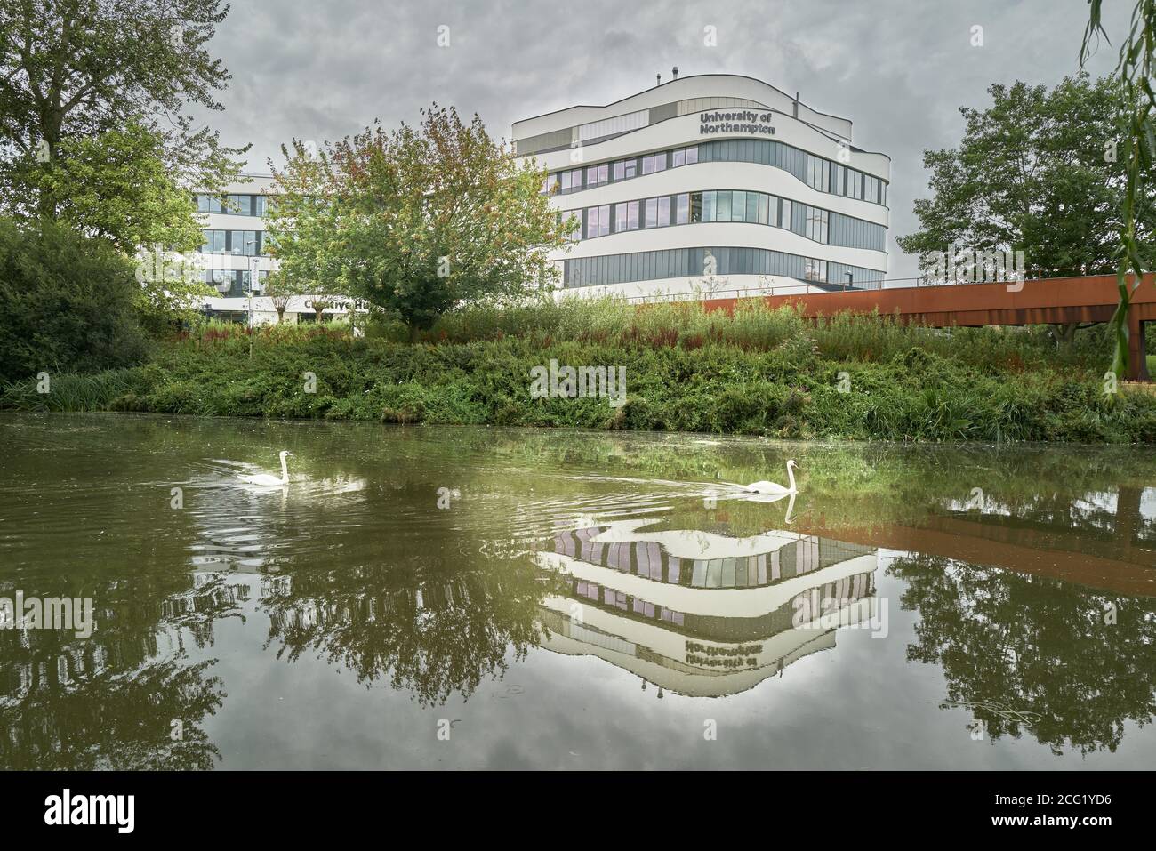 Un paio di cigni sorvola una riflessione sul fiume Nene dell'edificio Creative Hub presso il campus Riverside dell'Università di Northampton (UON), Foto Stock