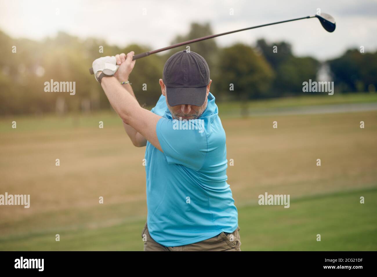 Uomo che gioca a golf oscillando alla palla come gioca il suo scatto utilizzando un driver visto da dietro guardando verso il basso il fairway in un sano stile di vita attivo co Foto Stock