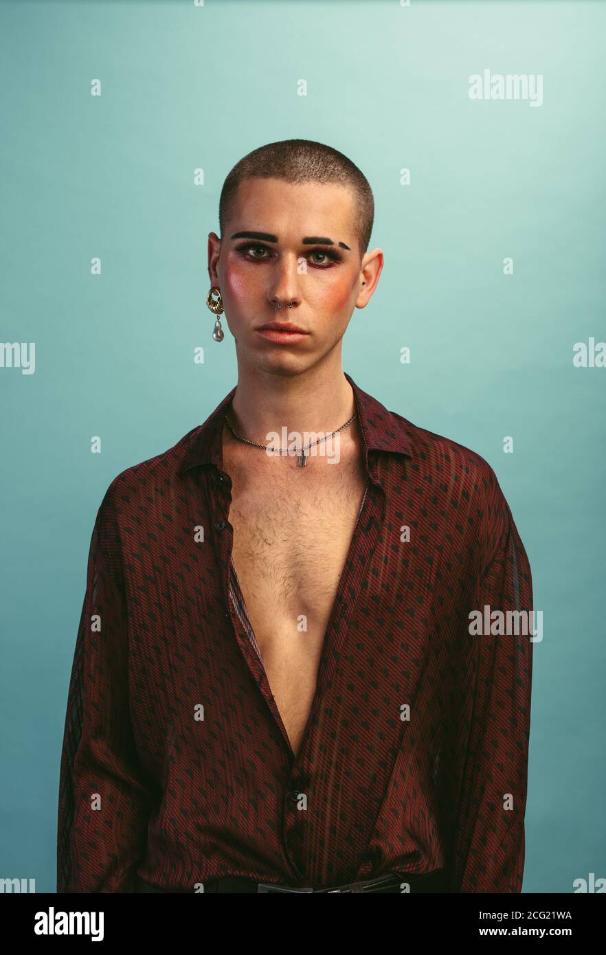 Giovane uomo con il trucco delle donne in piedi in studio con espressione vuota. Sesso fluido maschio su sfondo blu. Foto Stock
