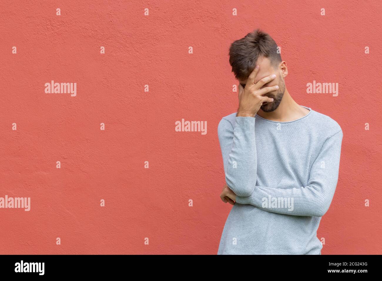 Facepalm. Vergognato uomo imbarazzato coprendo il viso. Ragazzo giovane su sfondo rosso parete con spazio di copia. Concetto di espressione facciale e sensazioni emotive Foto Stock