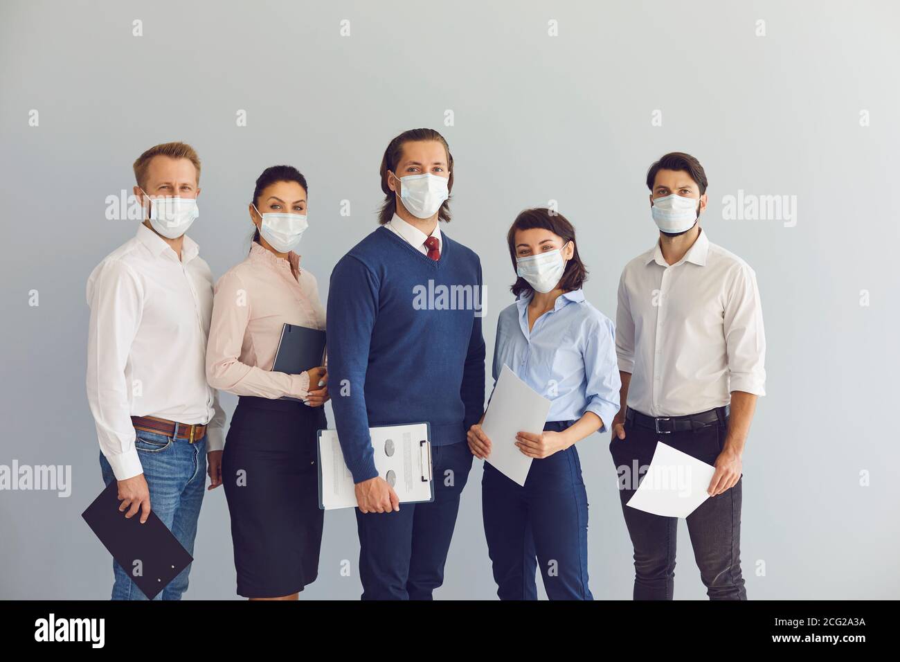 Gruppo di giovani uomini d'affari positivi in maschere di protezione medica in piedi con documenti in ufficio Foto Stock