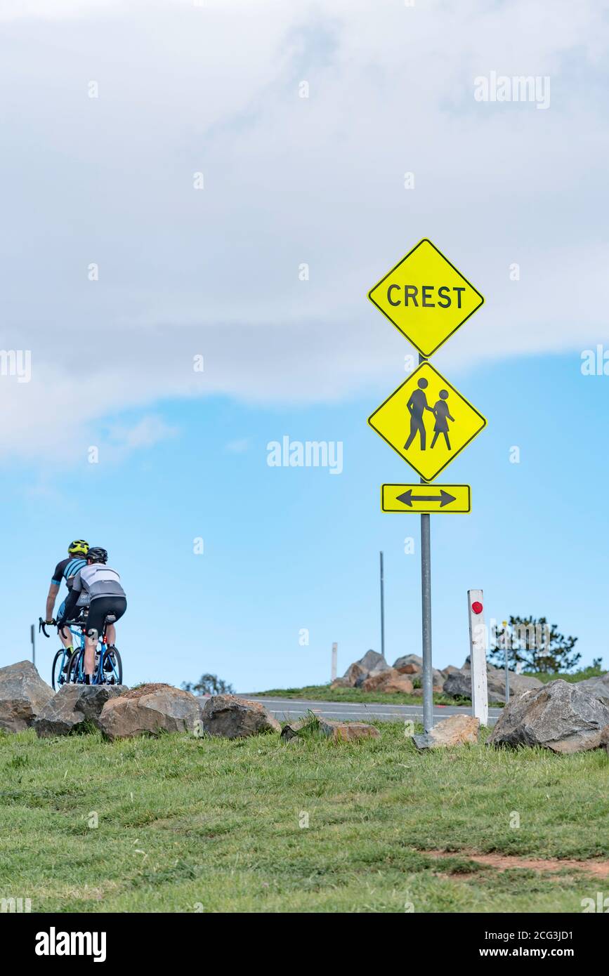 Due ciclisti maschi si dirigono verso la cresta di una collina, oltrepassando un cartello Crest su Dairy Farmers Hill a Canberra, ACT, Australia Foto Stock