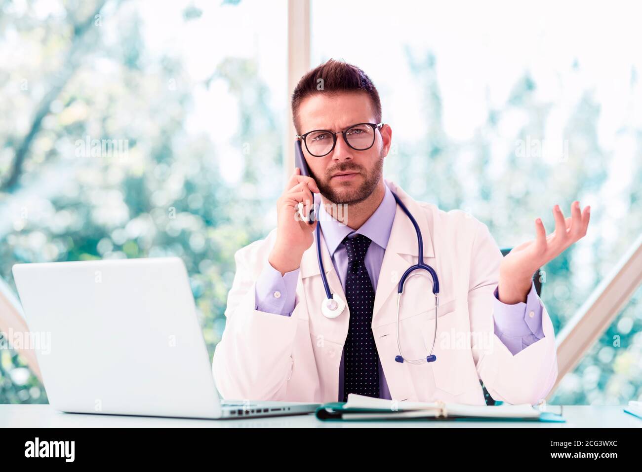 Un colpo di medico maschile seduto alla scrivania dietro il suo laptop e che parla con il suo paziente sul cellulare. Foto Stock