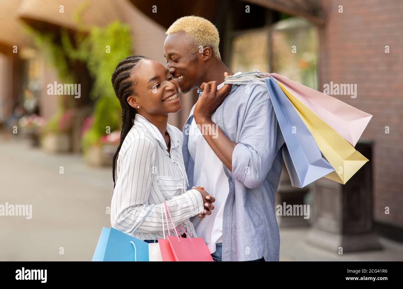 Amore, tempo libero e shopping. Allegro ragazzo afroamericano bacia la ragazza con borse colorate Foto Stock