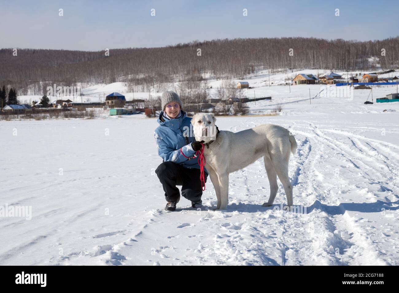 Una giovane ragazza è seduta accanto ad un grande cane da pastore bianco su un lago ghiacciato di neve sullo sfondo del villaggio. Foto Stock