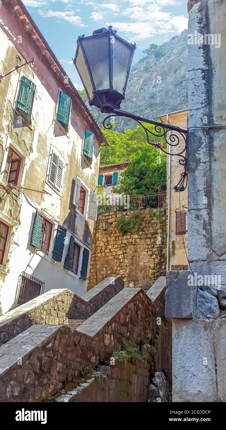 Stretta scala in pietra tra vecchi edifici con persiane in legno nel centro storico di Kotor, con le montagne Lovcen e il cielo blu sullo sfondo. Foto Stock