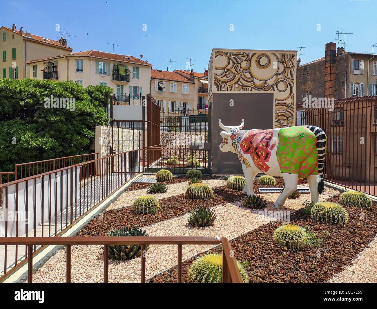 Vista del cortile con edifici francesi tradizionali con tetti in tegole rosse, cactus a forma di palla in piccole pietre e colorato scultura di mucca dipinto. Foto Stock