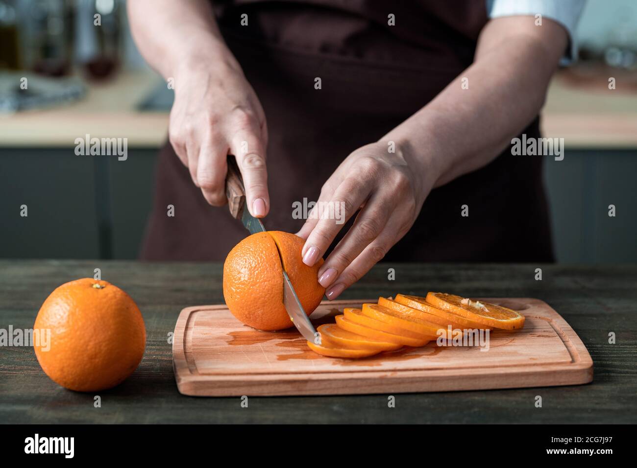 Primo piano di una chef femminile irriconoscibile in grembiule in piedi al bancone e tagliando arancio su tavola di legno Foto Stock