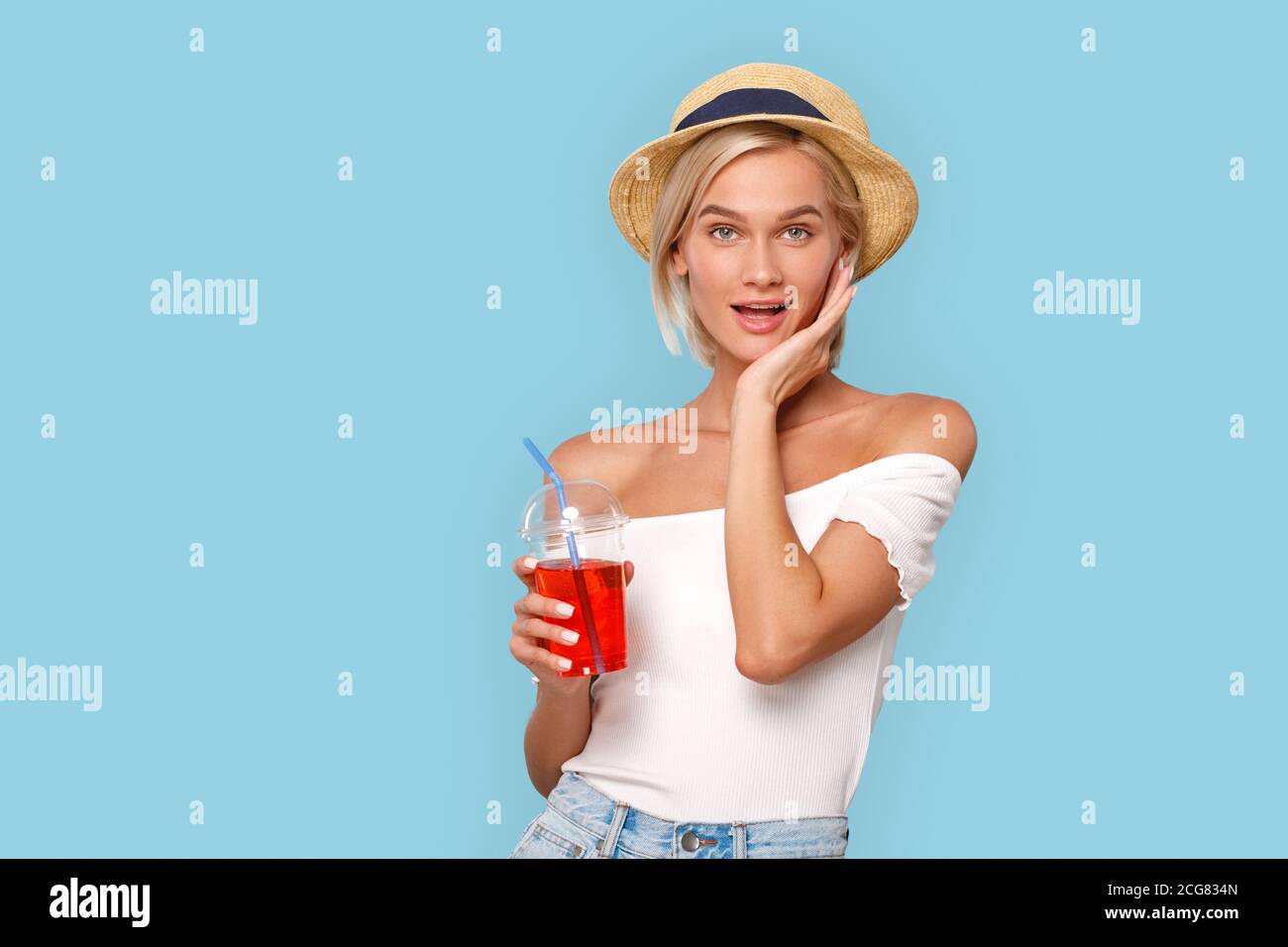Stile libero. Giovane bionda donna in cima e cappello isolato su blu con una tazza di succo in posa per la macchina fotografica giocoso Foto Stock