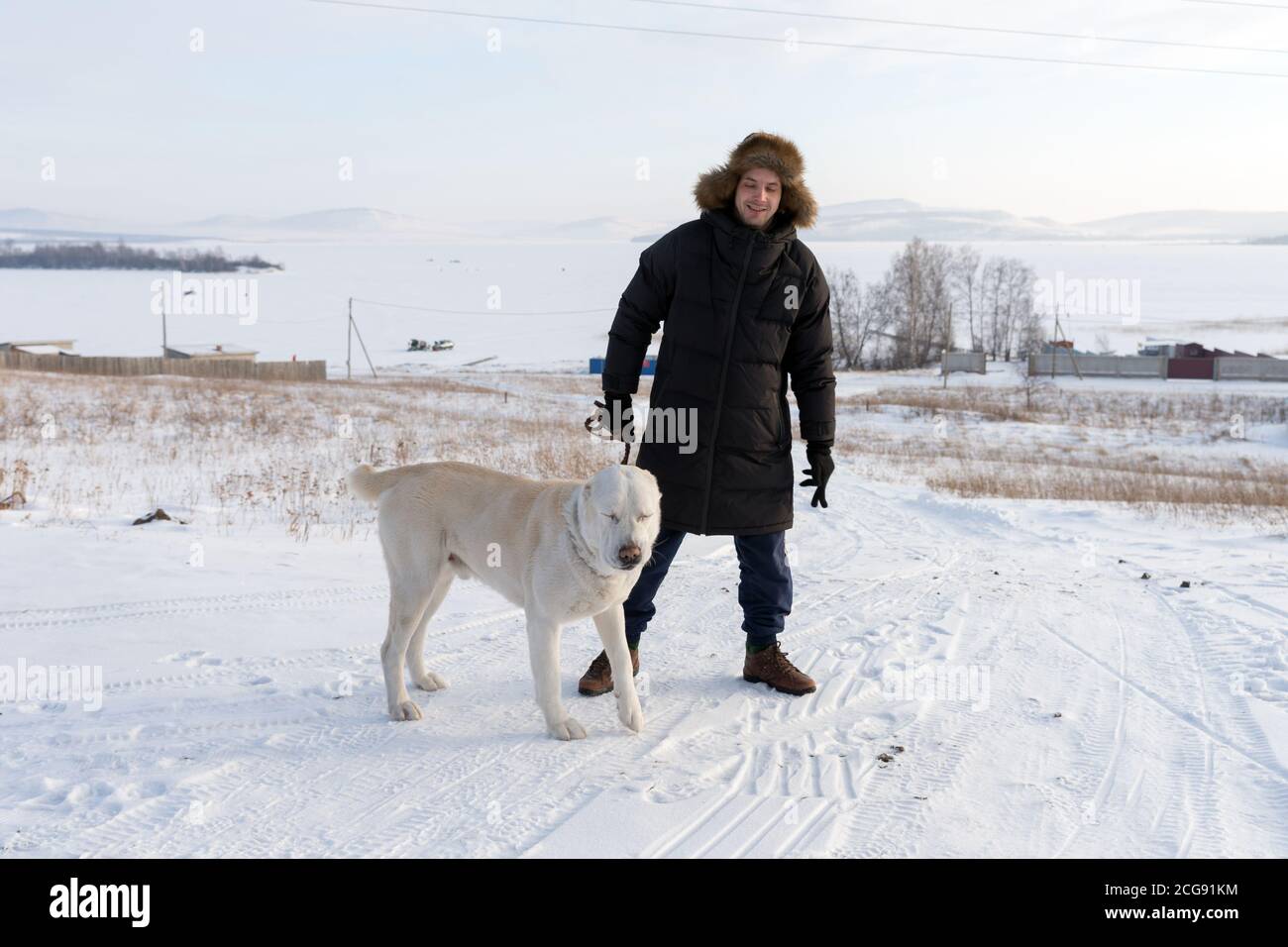 Un giovane si alza con un cane grande su una strada rurale in mezzo a un lago ghiacciato in inverno. Foto Stock