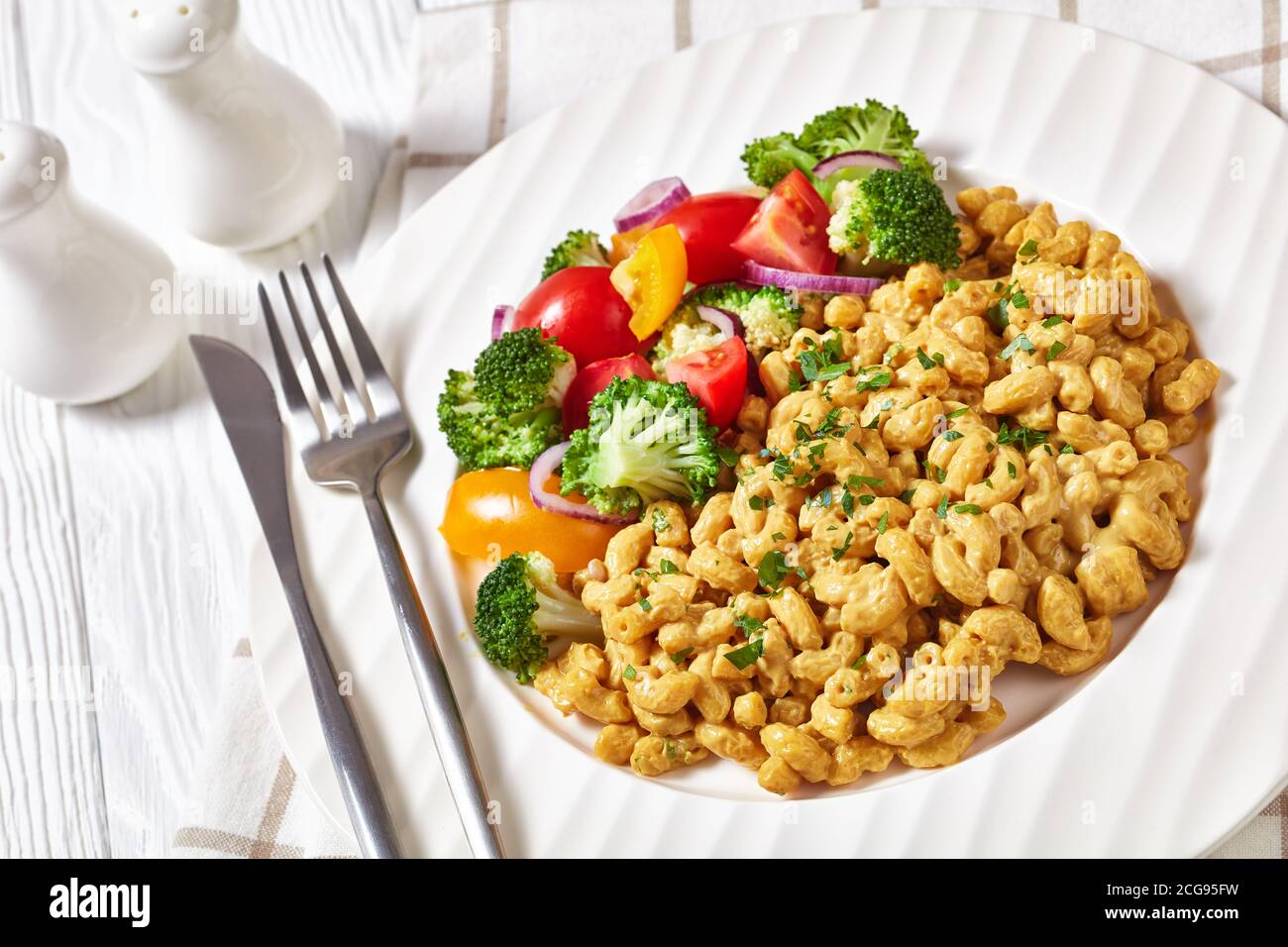 mac vegan e formaggio con salsa di lievito nutrizionale e insalata di verdure su un piatto bianco su un tavolo di legno bianco con posate in argento, vista orizzontale fr Foto Stock