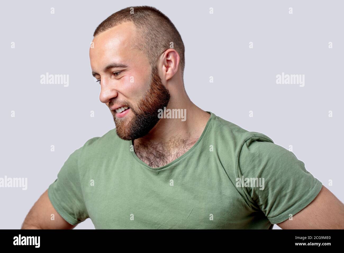 Uomo caucasico dal bearded atletico in t-shirt casual kaki su sfondo bianco in umore pensivo . Ritratto di giovane sorridente contro la parete grigia. Foto Stock