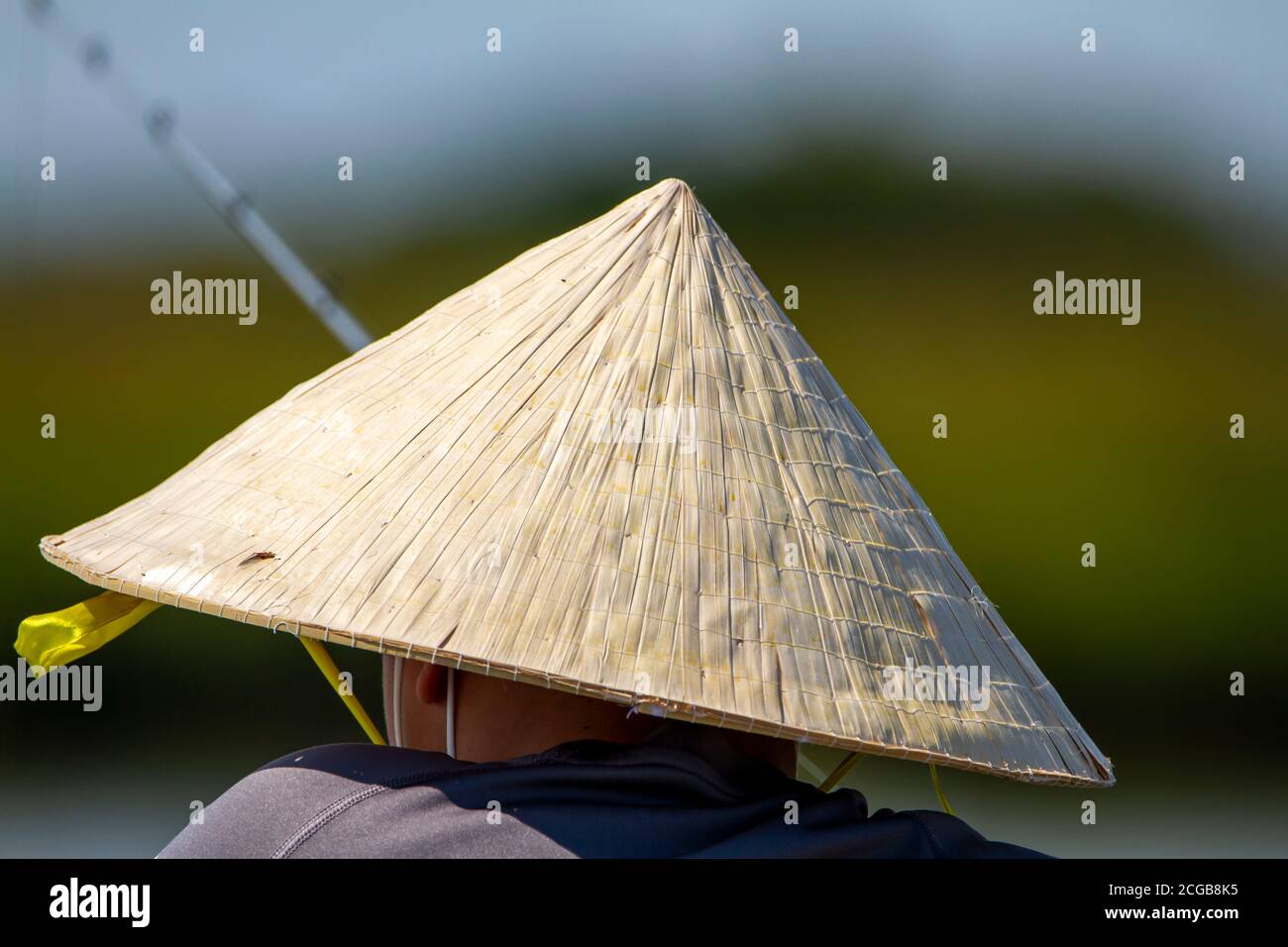 Un uomo asiatico che indossa un tradizionale cappello di bambù di paglia è in piedi vicino alle barriere di legno di un ponte presso un lago e la pesca con una canna da pesca. Foto Stock
