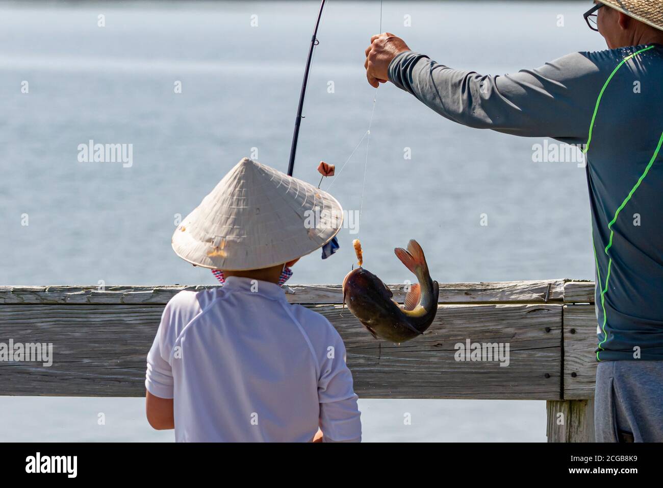 Un ragazzo asiatico che indossa tradizionale bambù cinese cool paglia cappello conico è la pesca con suo padre su un ponte. Il ragazzo ha appena catturato un pesce gatto un Foto Stock
