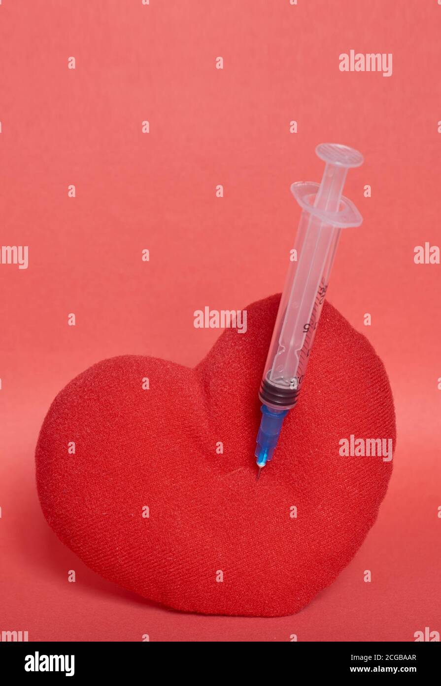 Un cuore giocattolo imbottito con una siringa bloccata in esso. Sfondo rosso. Formato verticale Foto Stock