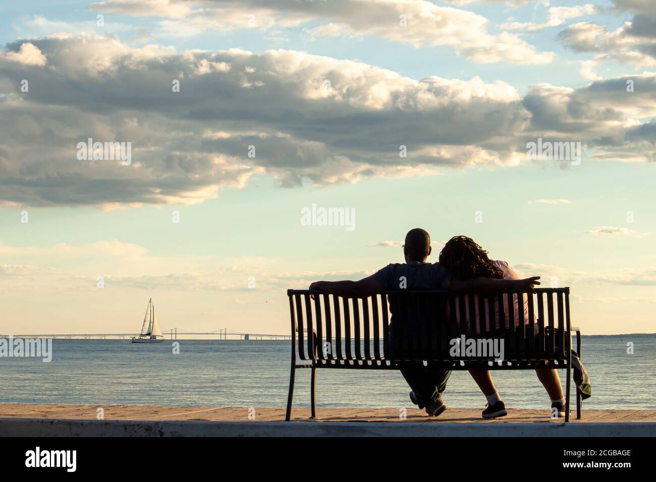Una giovane coppia afroamericana è seduta su una panchina che guarda al mare e al ponte di Chesapeake Bay. L'uomo mette il suo braccio sopra la spalla della donna w Foto Stock