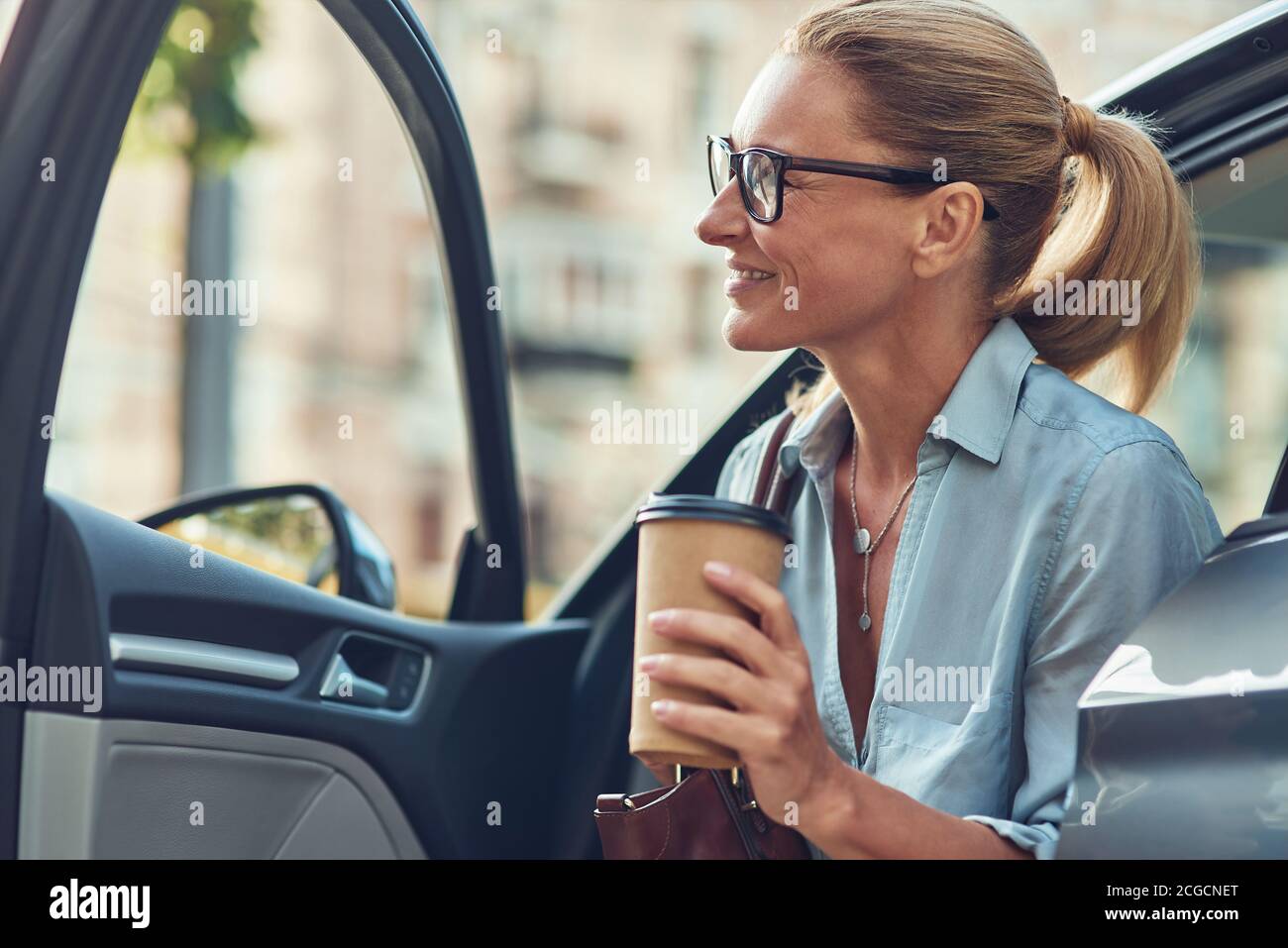 Felice riuscito metà anziana donna di affari che tiene la tazza di carta del caffè mentre esce dalla sua automobile, sta arrivando al lavoro Foto Stock