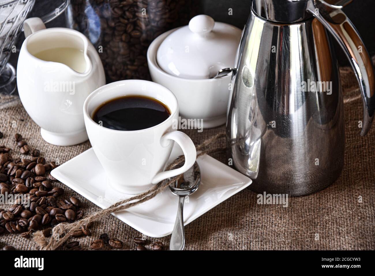 una tazza di caffè con piatti bianchi, una caraffa di latte e una pentola di zucchero serviti a juta con thermos d'argento Foto Stock