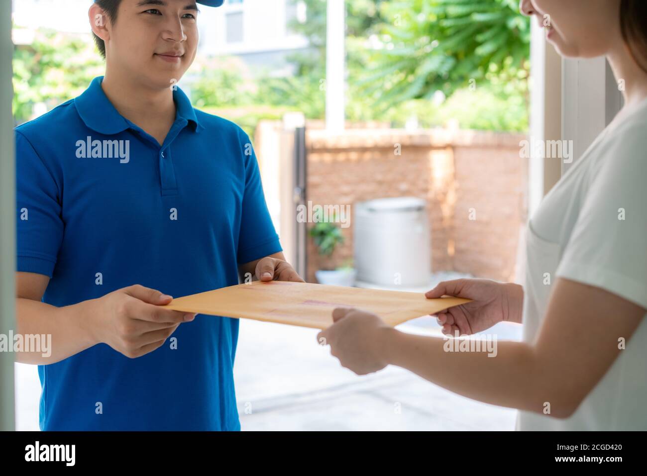 Il giovane uomo di consegna asiatico in sorriso uniforme blu e tenendo la lettera o la busta del documento nella casa anteriore e la donna asiatica che accetta una consegna della busta Foto Stock