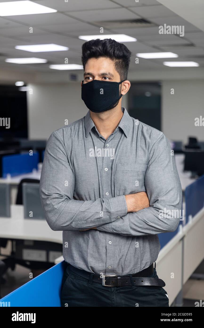 Ritratto di bel giovane uomo d'affari indiano indossare maschera di protezione Covid in piedi in ufficio, ambiente aziendale, in piedi con le braccia incrociate. Foto Stock