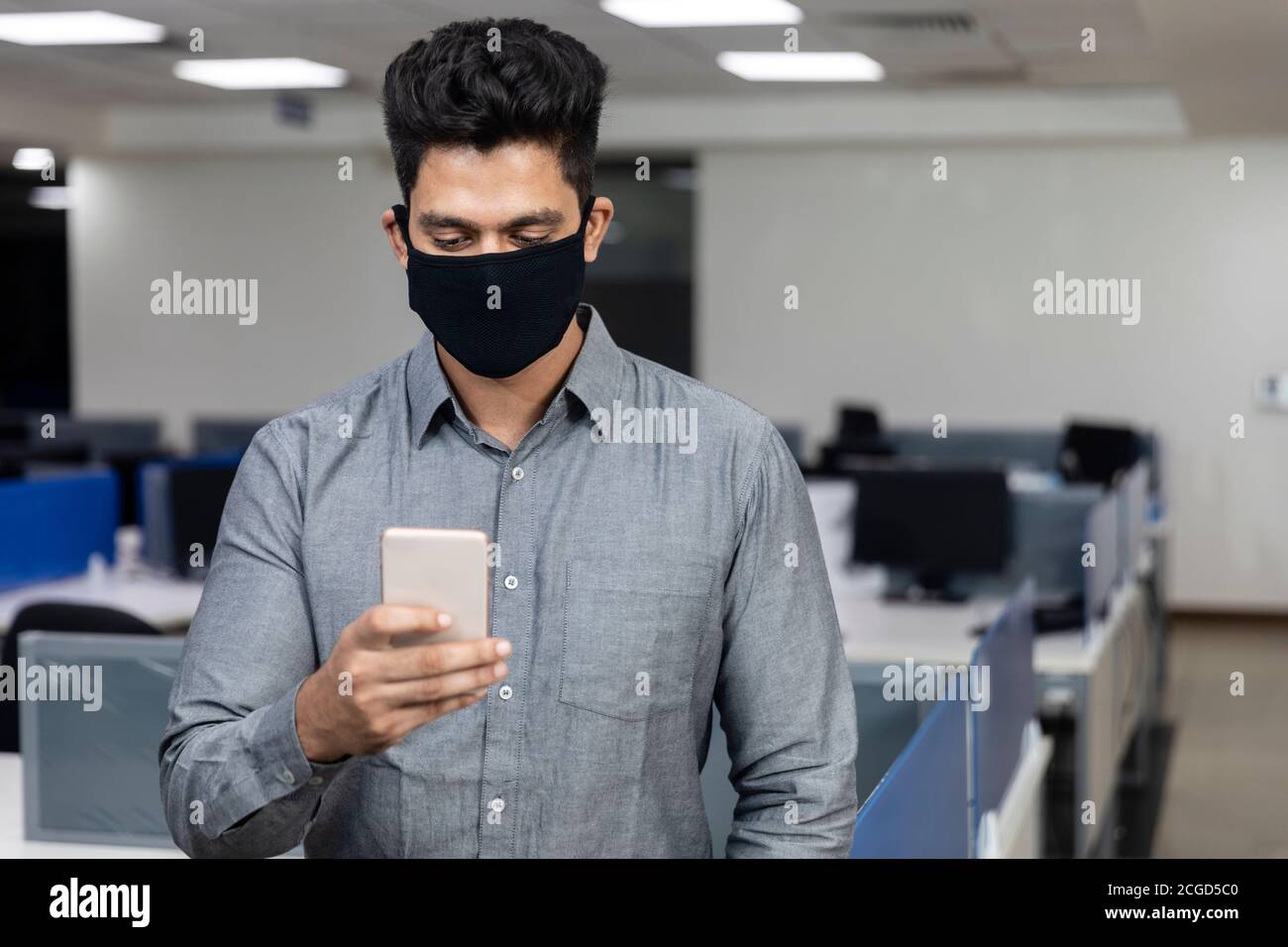 Ritratto di bel giovane uomo d'affari indiano indossare maschera di protezione Covid e controllare il suo telefono in un ambiente aziendale. Foto Stock