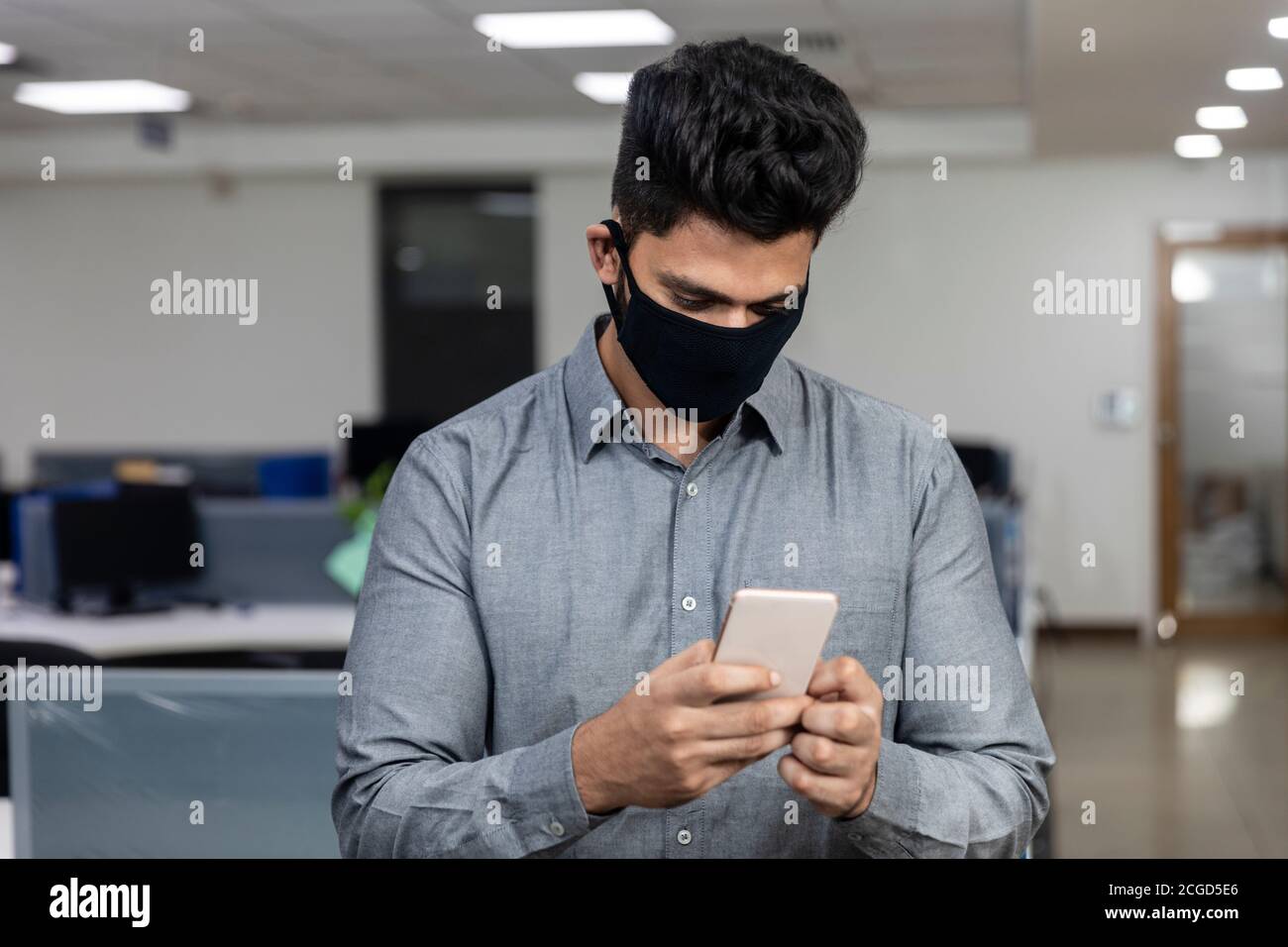 Ritratto di bel giovane uomo d'affari indiano indossare maschera di protezione Covid e controllare il suo telefono in un ambiente aziendale. Foto Stock