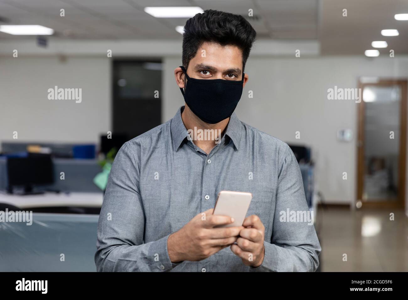 Ritratto di bel giovane uomo d'affari indiano indossare maschera di protezione Covid e controllare il suo telefono in un ambiente aziendale. Foto Stock