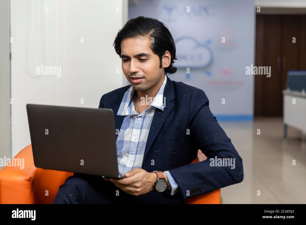 Ritratto di un giovane uomo d'affari indiano di successo in tuta oscura seduto sul divano e lavorando su un computer portatile, ambiente aziendale. Foto Stock