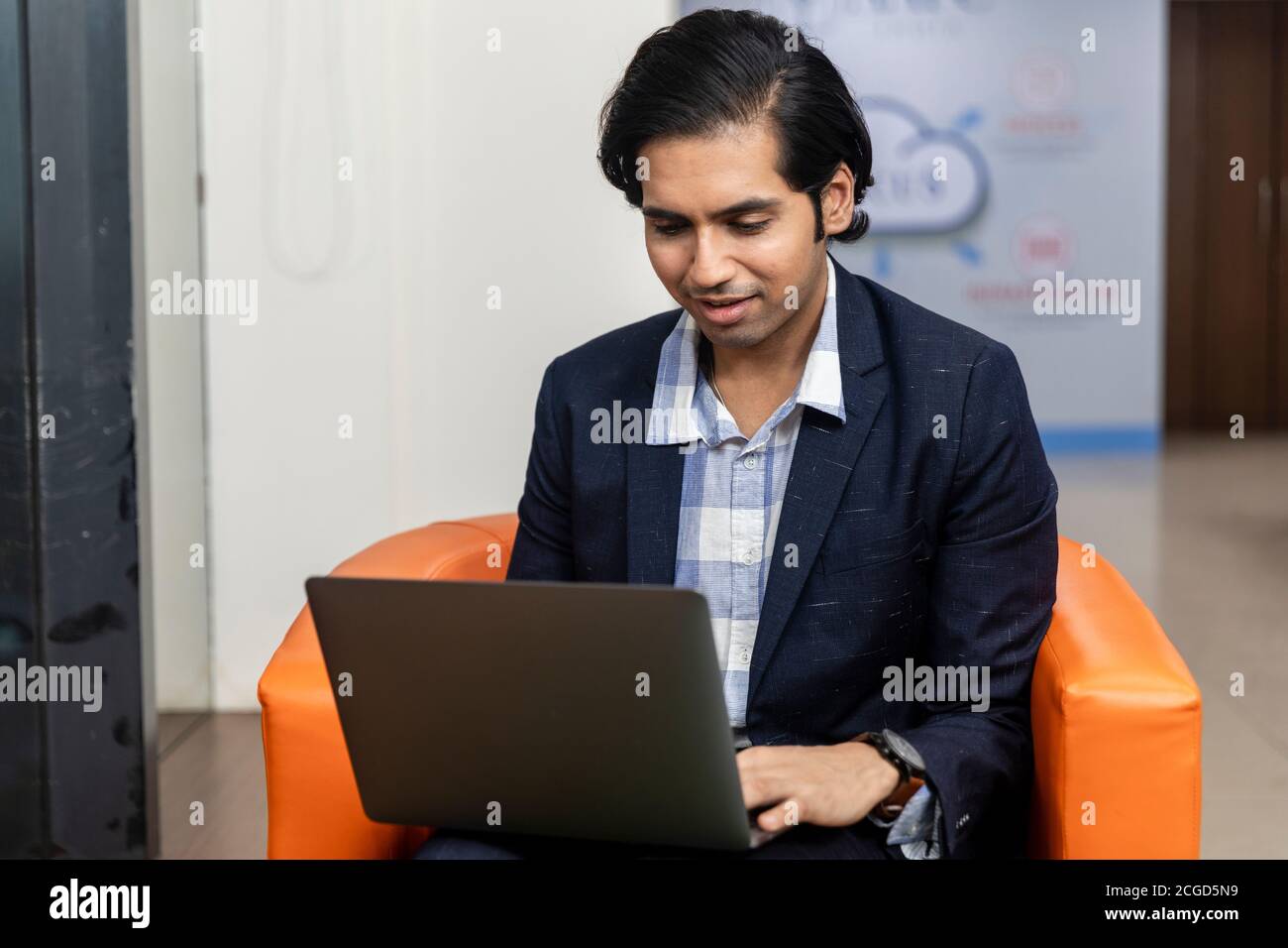 Ritratto di un giovane uomo d'affari indiano di successo in tuta oscura seduto sul divano e lavorando su un computer portatile, ambiente aziendale. Foto Stock