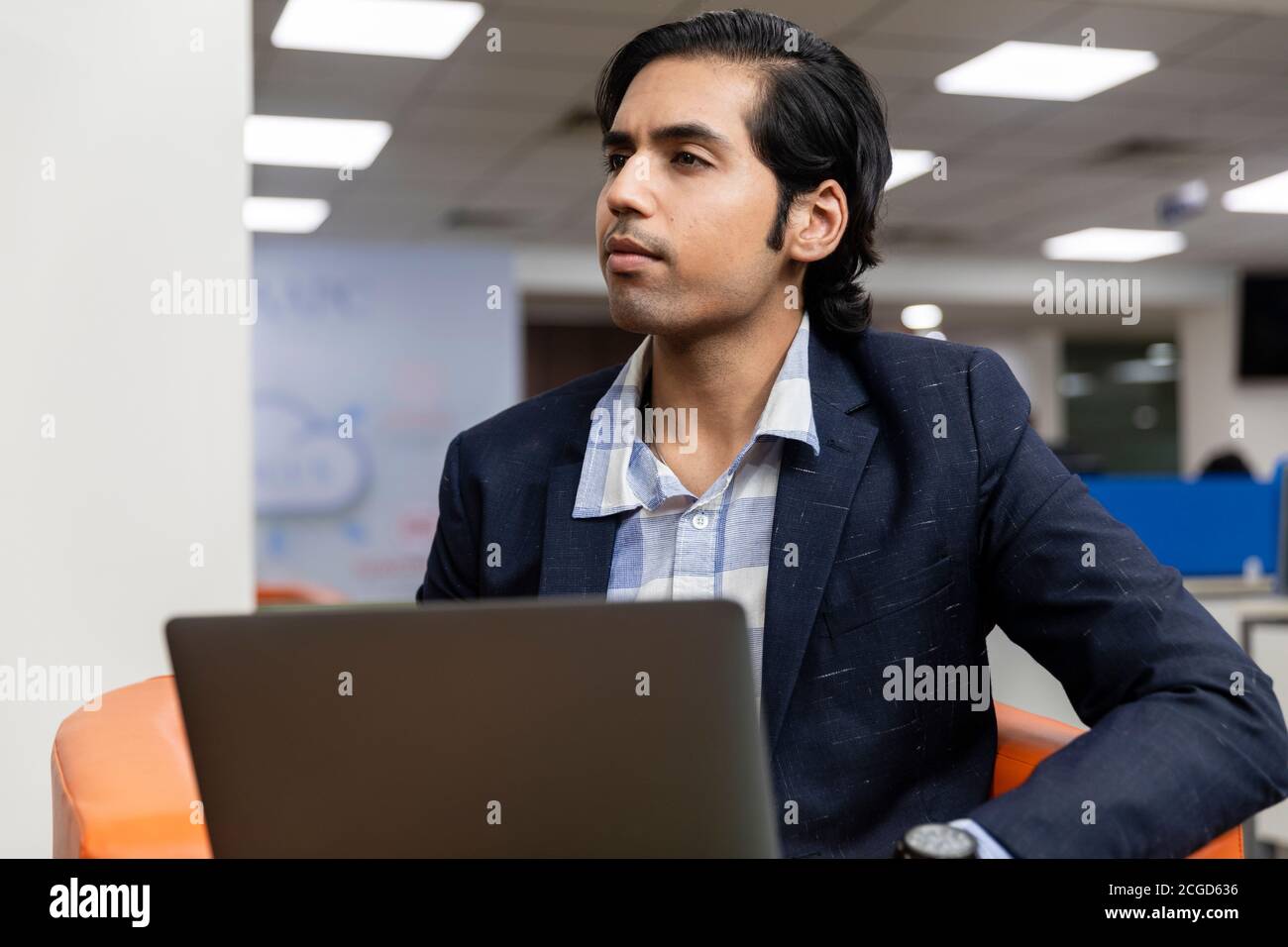 Ritratto di un giovane uomo d'affari indiano di successo in tuta oscura seduto sul divano e lavorando su un computer portatile, ambiente aziendale. Foto Stock