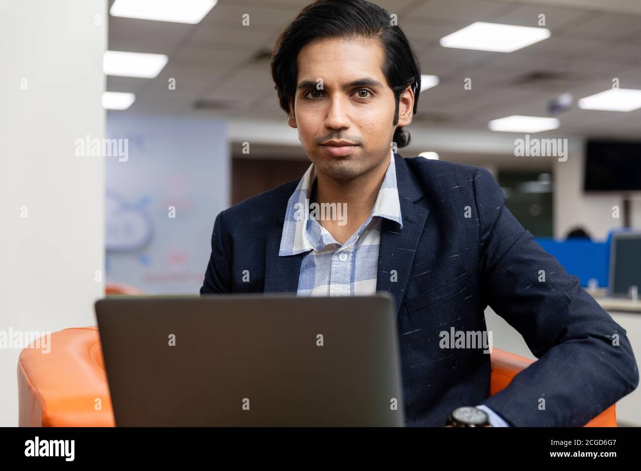 Ritratto di un giovane uomo d'affari indiano di successo in tuta oscura seduto sul divano e lavorando su un computer portatile, ambiente aziendale. Foto Stock