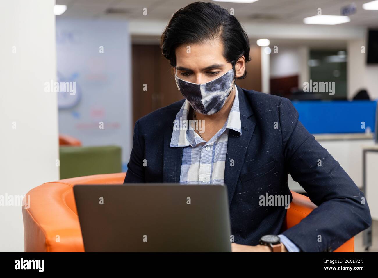 Ritratto di giovane uomo d'affari indiano di successo in abito scuro indossando maschera di protezione Covid e lavorando su laptop, ambiente aziendale. Foto Stock
