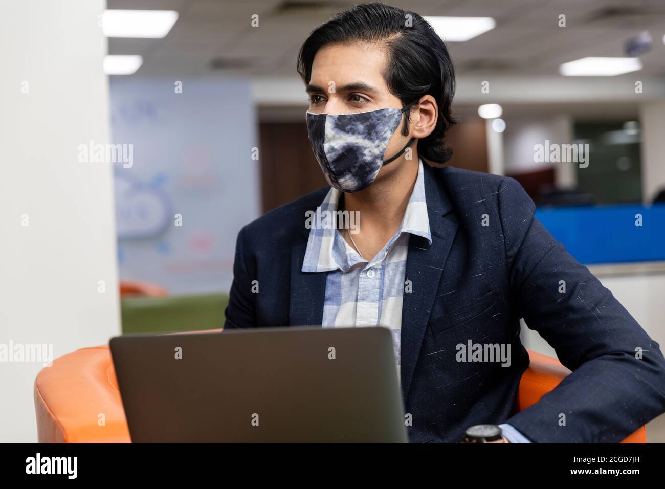 Ritratto di giovane uomo d'affari indiano di successo in abito scuro indossando maschera di protezione Covid e lavorando su laptop, ambiente aziendale. Foto Stock
