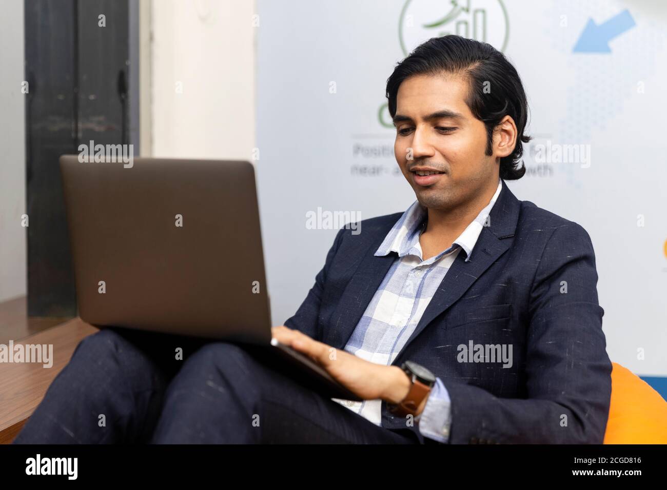 Ritratto di un giovane uomo d'affari indiano di successo in tuta oscura seduto e lavorando su laptop, ambiente aziendale, volto espressivo. Foto Stock