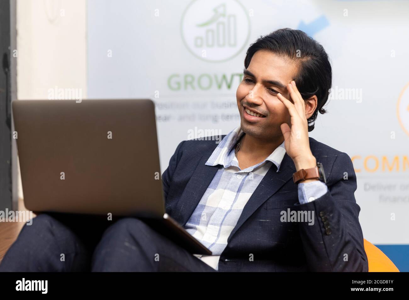 Ritratto di un giovane uomo d'affari indiano di successo in tuta oscura seduto e lavorando su laptop, ambiente aziendale, volto espressivo. Foto Stock