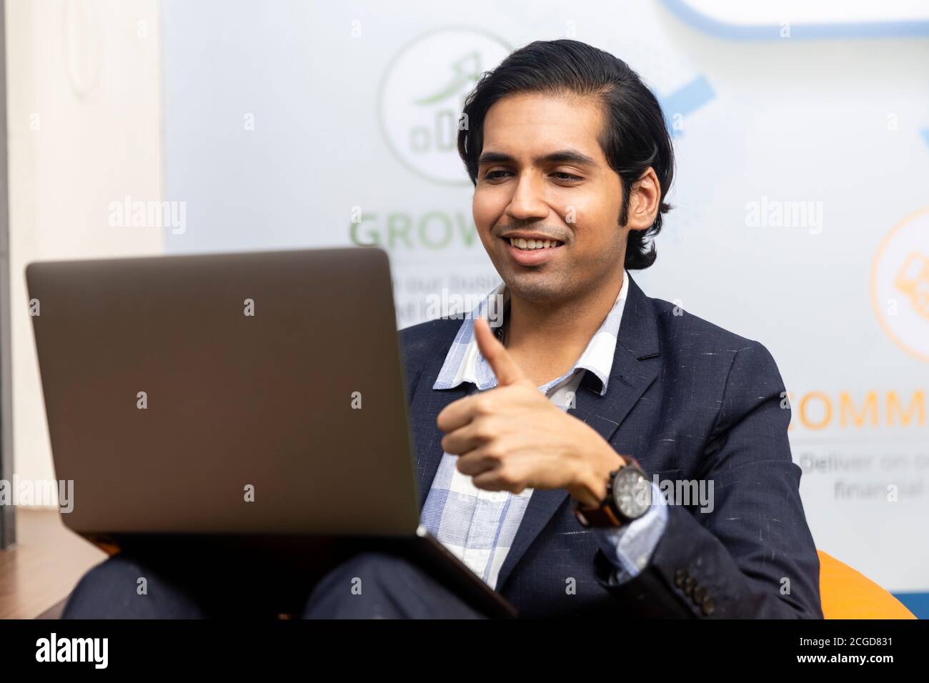 Ritratto di un giovane uomo d'affari indiano di successo in tuta oscura seduto e lavorando su laptop, ambiente aziendale, volto espressivo. Foto Stock