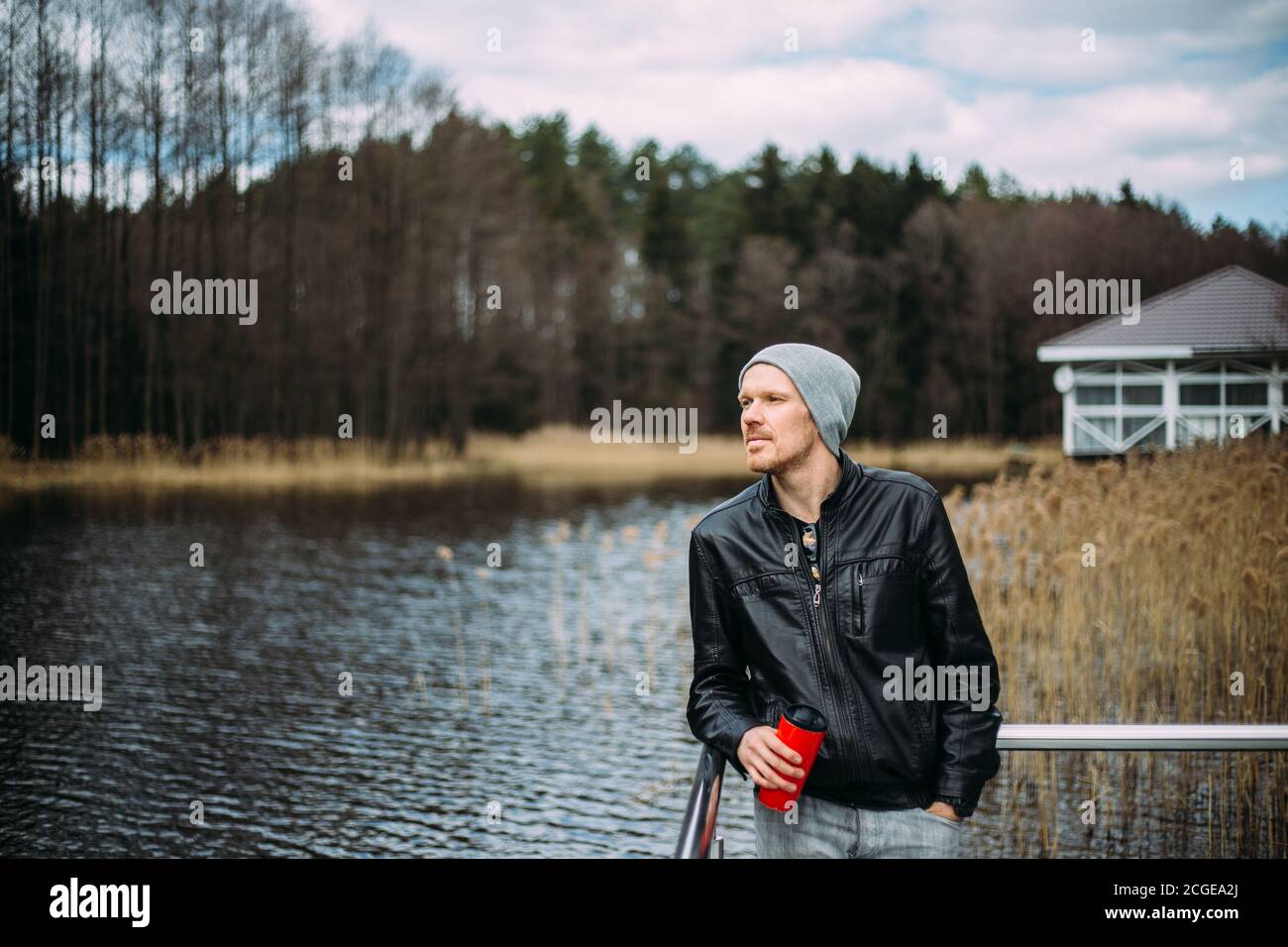 Uomo pusivo in una giacca di pelle e cappello si alza e pensa all'aperto. Buon modello maschile guardando pensivo al lago. Foto Stock