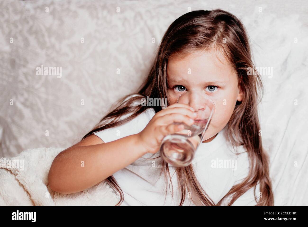 un bambino malato, una ragazza seduta su un letto con un cuscino bianco, bevendo acqua da un bicchiere Foto Stock
