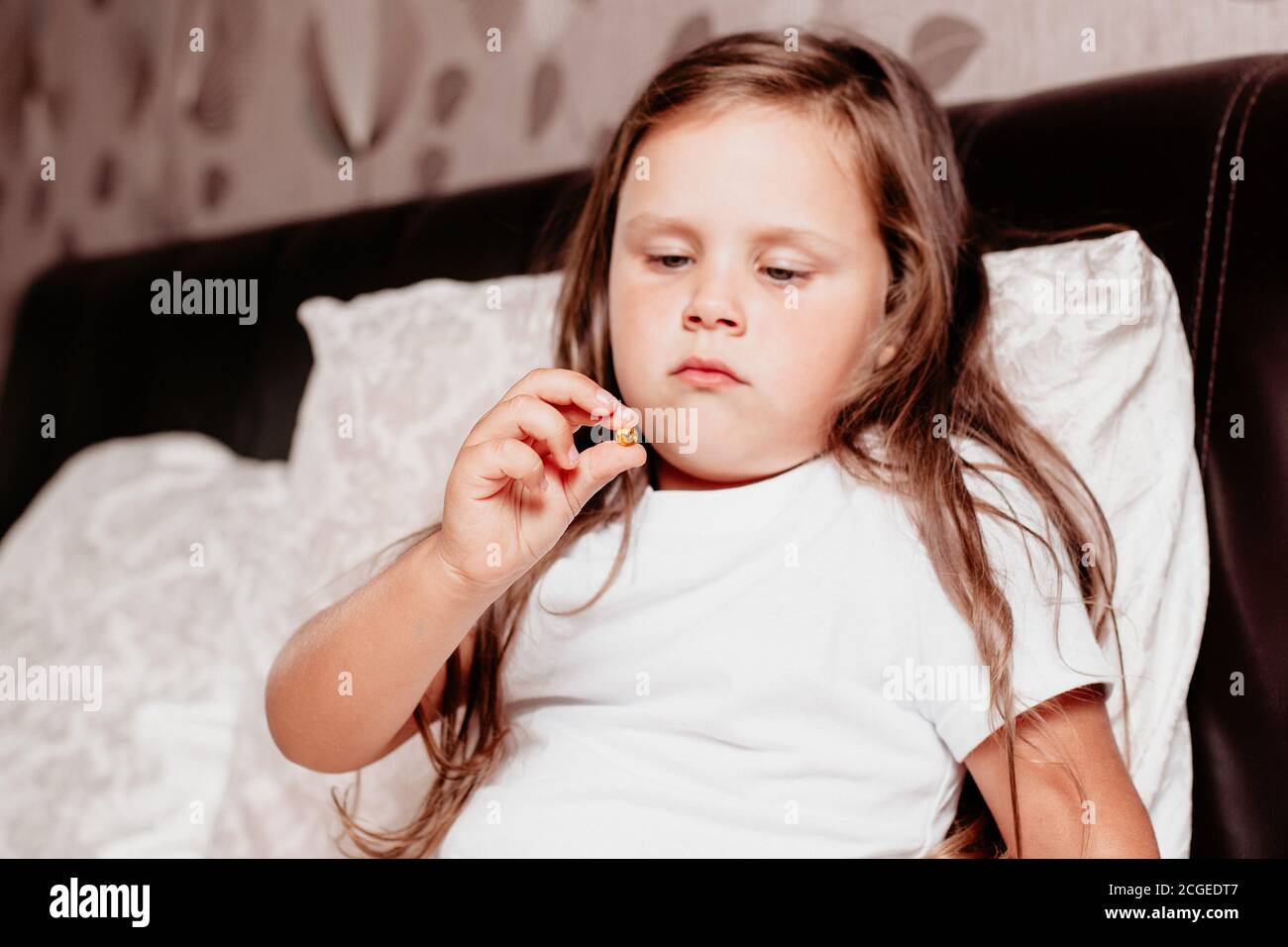 un bambino malato, una ragazza seduta su un letto con un cuscino bianco, guardando una vitamina in mano Foto Stock