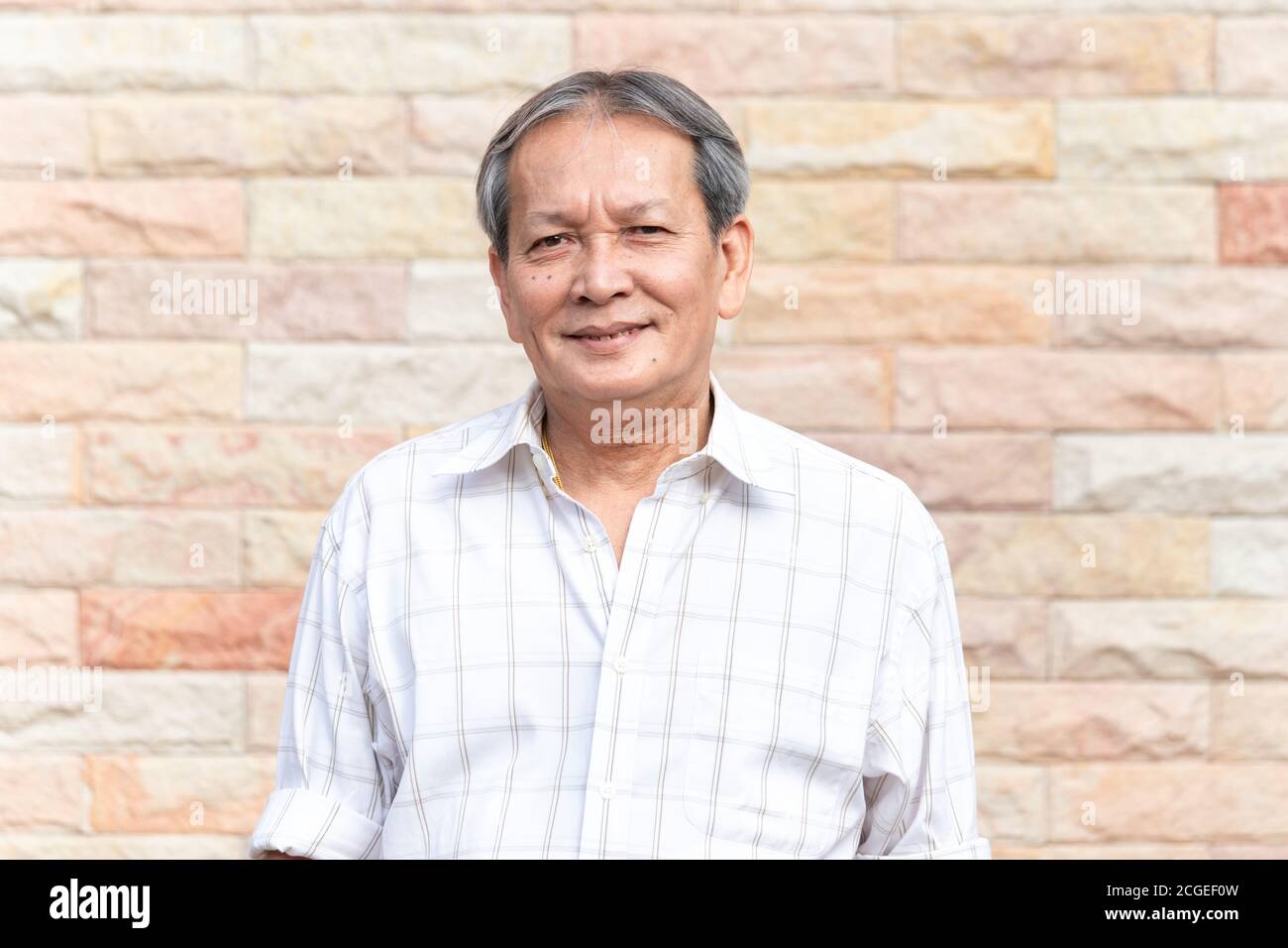 Ritratto di sorridente anziano asiatico uomo in piedi di fronte a muro di mattoni sfondo con modo sicuro di sé. Primo piano del sorriso più anziano con i capelli grigi Foto Stock