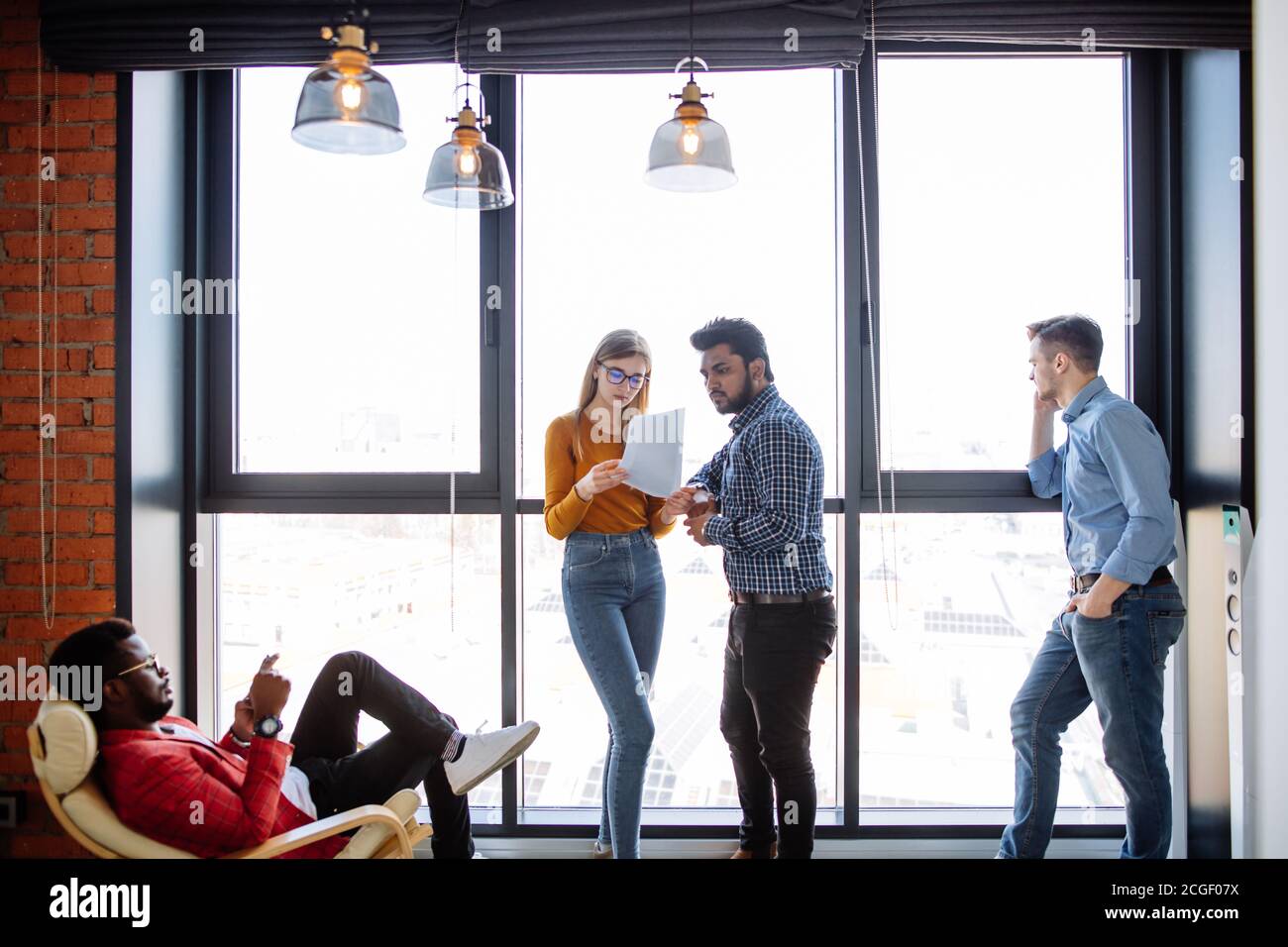 Gruppo multietnico di studenti che si aggirano nella sala universitaria durante la recinita, in piedi accanto alla finestra che parla o comunica nei social media tramite un computer portatile Foto Stock