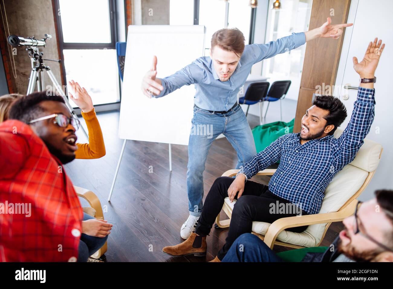 Gruppo di giovani entusiasti che sollevano le mani nello spirito di squadra alto cinque sopra il tavolo, seduti uno di fronte all'altro in uno spazio ben illuminato studio dietro Foto Stock