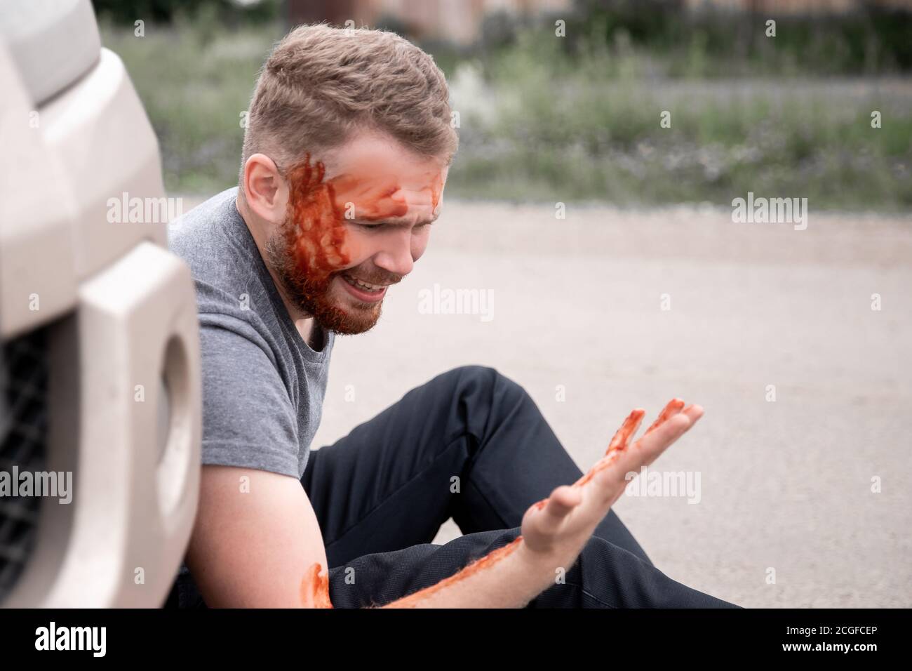 Concetto di assicurazione. Incidente d'auto, l'uomo si siede con la testa sanguinosa vicino alla ruota, gridando per aiuto in difficoltà Foto Stock