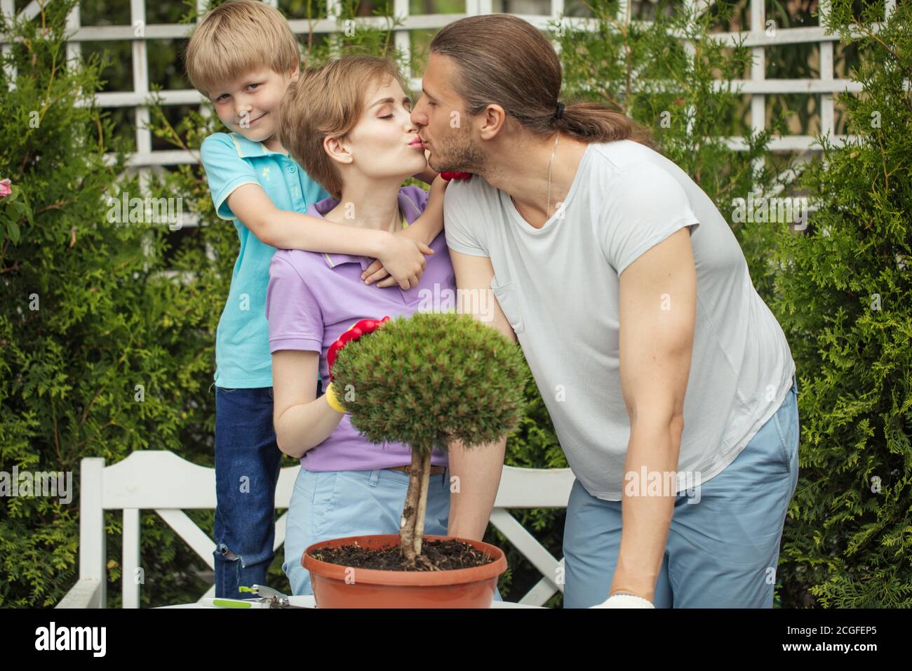 Ritratto di coppia sposata amorevole baciarsi su giardino verde abete-albero sfondo, e felice 6 anni figlio abbracciando Mum e guardando la macchina fotografica. Foto Stock