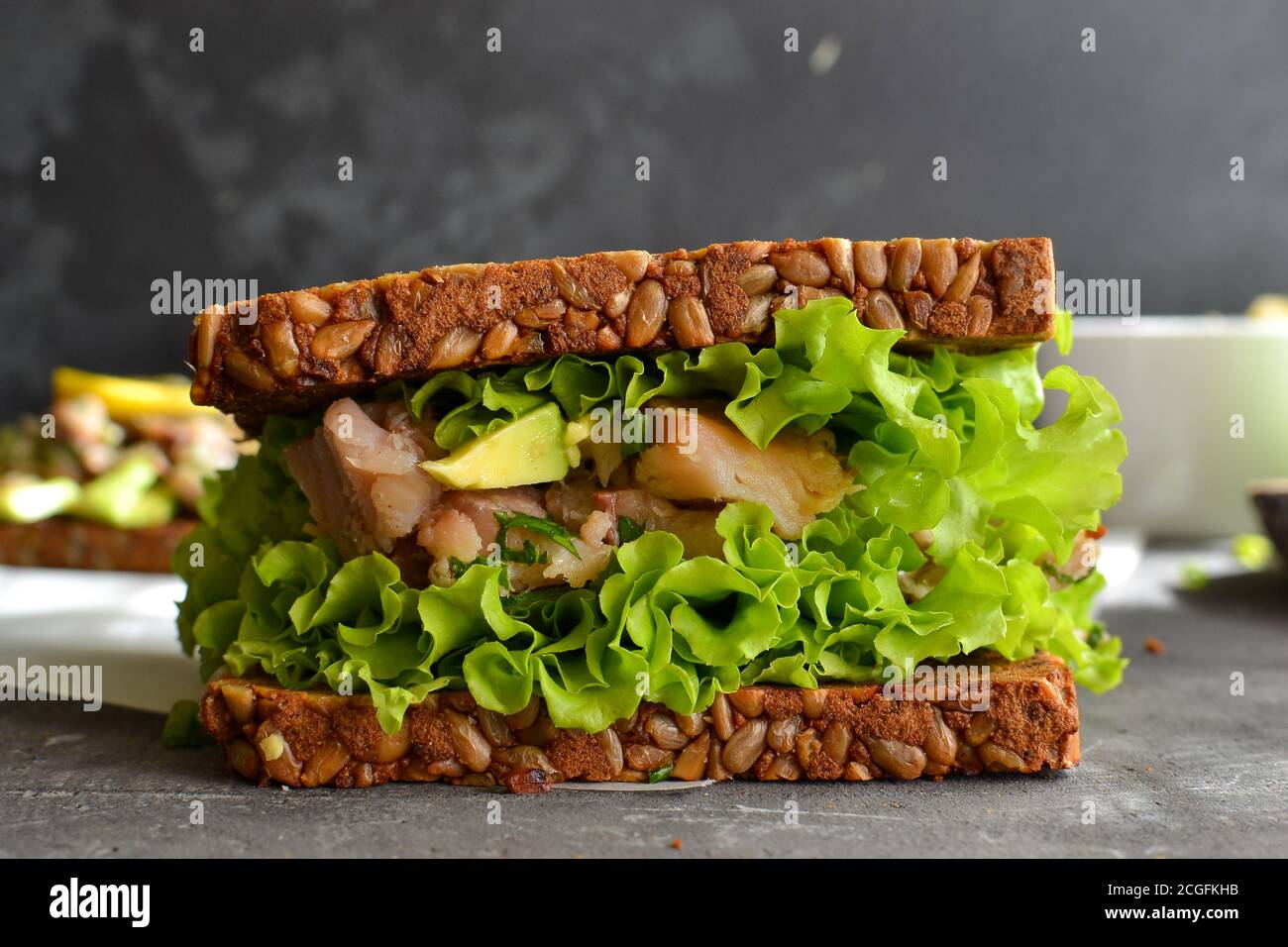 Panino scuro con semi. Sandwich con lattuga, avocado e pesce. Pranzo sano o brunch. Sfondo scuro. Primo piano. Foto Stock